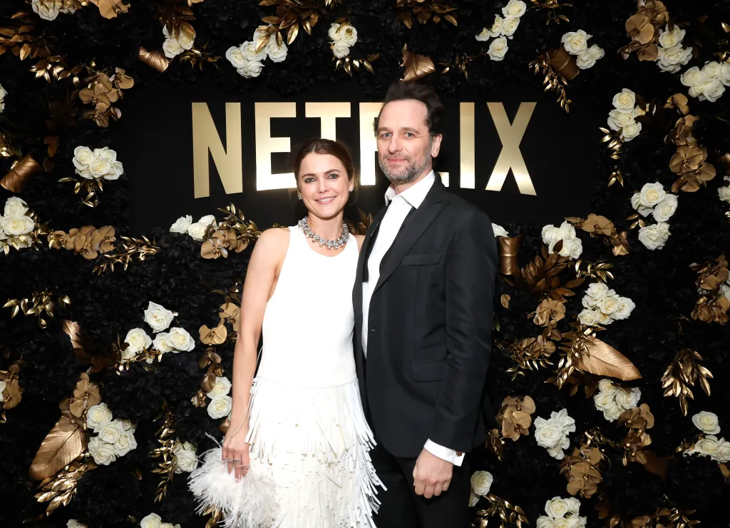 Keri Russell and Matthew Rhys pose in front of a black and gold Netflix backdrop adorned with white flowers.