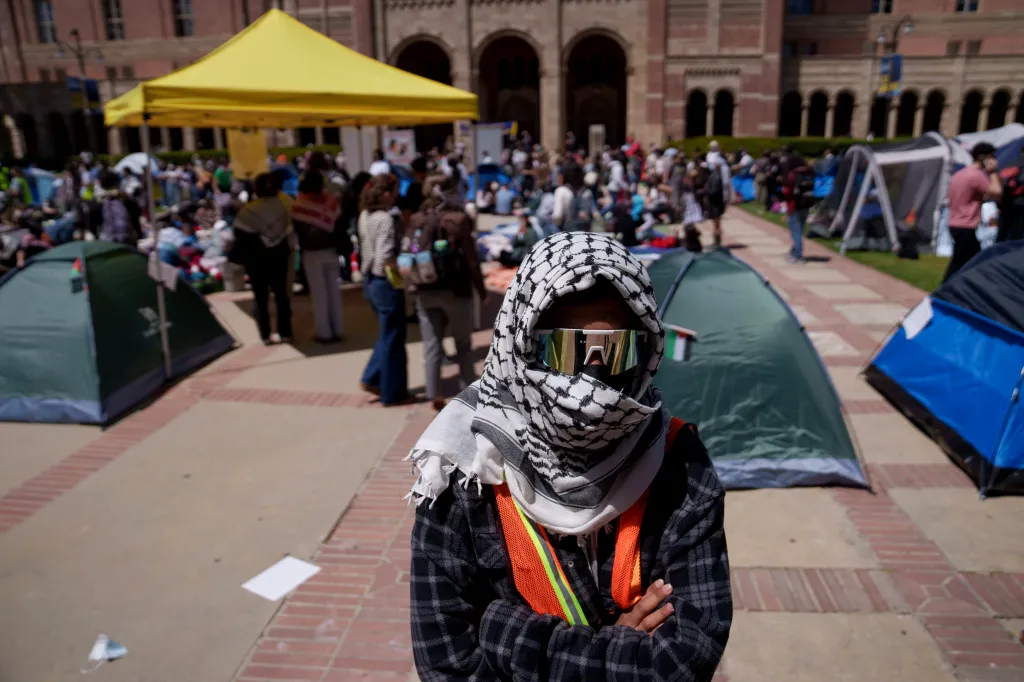 A pro-Palestinian demonstrator wearing a keffiyeh and sunglasses stands with arms crossed at a campus encampment.