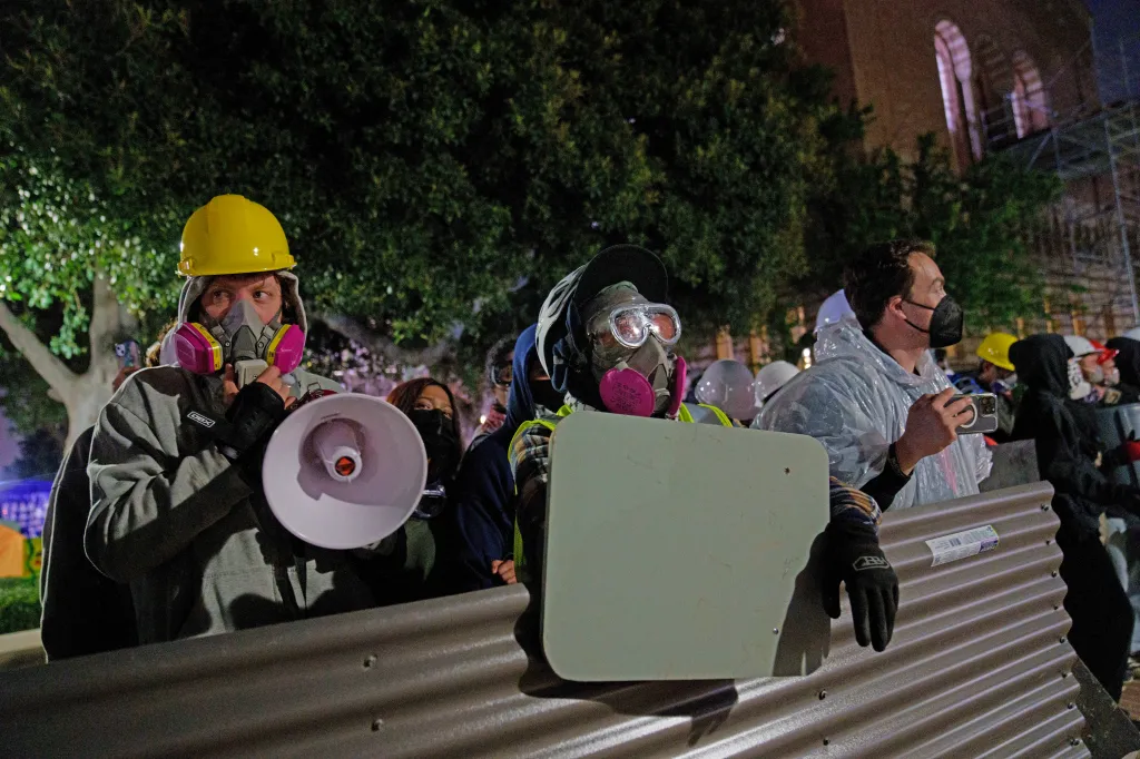 Pro-Palestine protestors wearing protective gear stand behind a metal barricade in an encampment at UCLA.