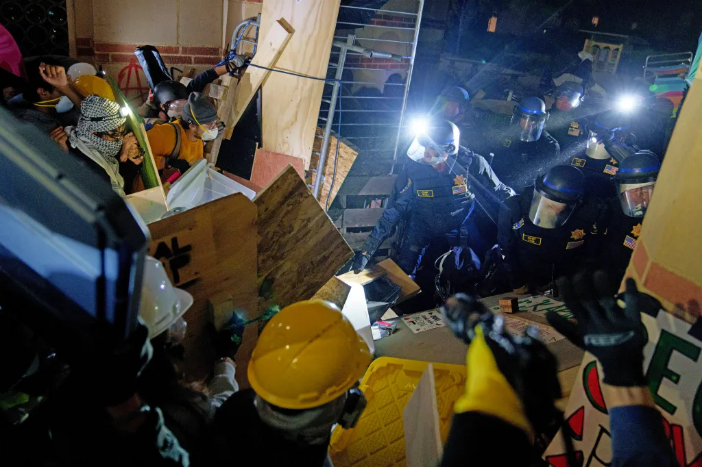 Law enforcement officers in riot gear break through wooden barricades set up by Pro-Palestinian protesters at an encampment.