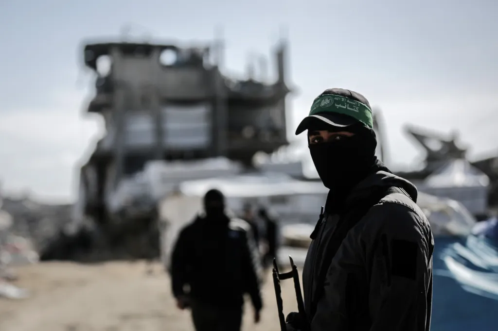 An armed militant from the al-Qassam Brigades wearing a face mask and a green headband stands in front of a ruined building in the Jabalia refugee camp.