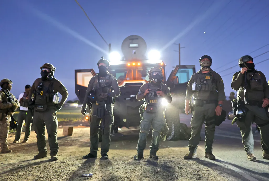 Federal agents in tactical gear stand in front of a law enforcement vehicle, blocking protesters outside a cannabis farm.
