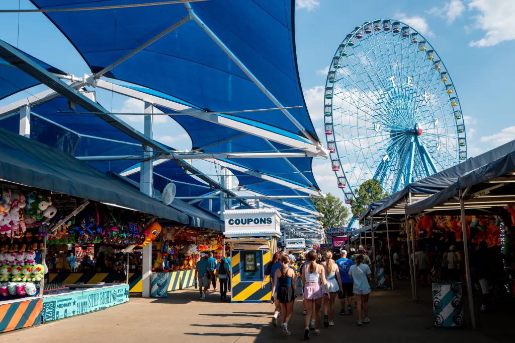 People walk along the midway at the State Fair of Texas, passing by carnival game booths and under large blue shade sails, with a large blue Ferris wheel in the background.