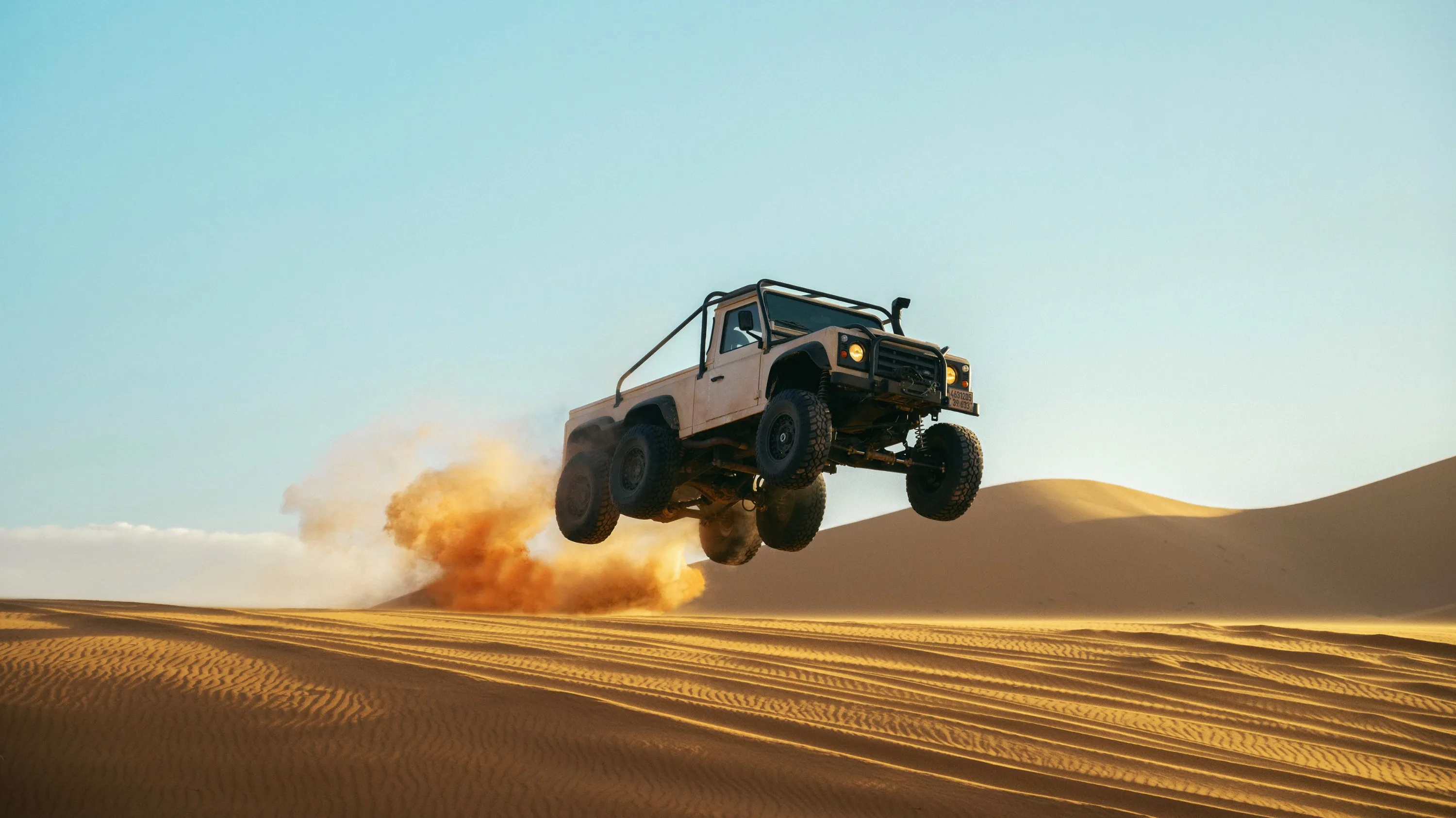 A beige off-road pickup truck leaps through the air over a desert landscape, kicking up a cloud of dust.