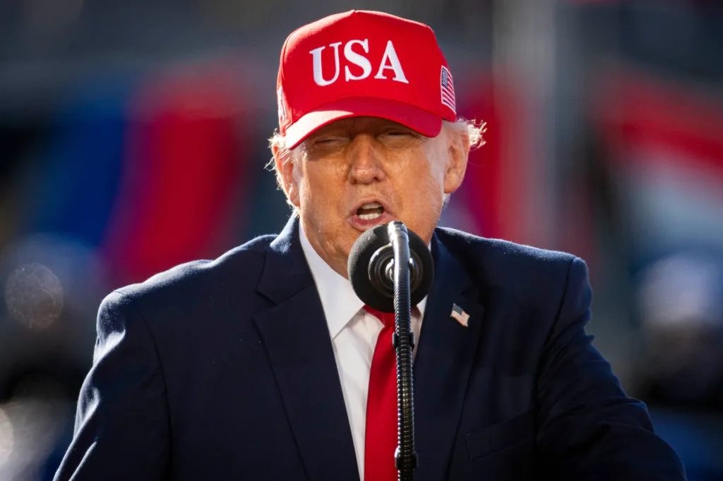 President Donald Trump wearing a red USA hat and speaking at a podium.
