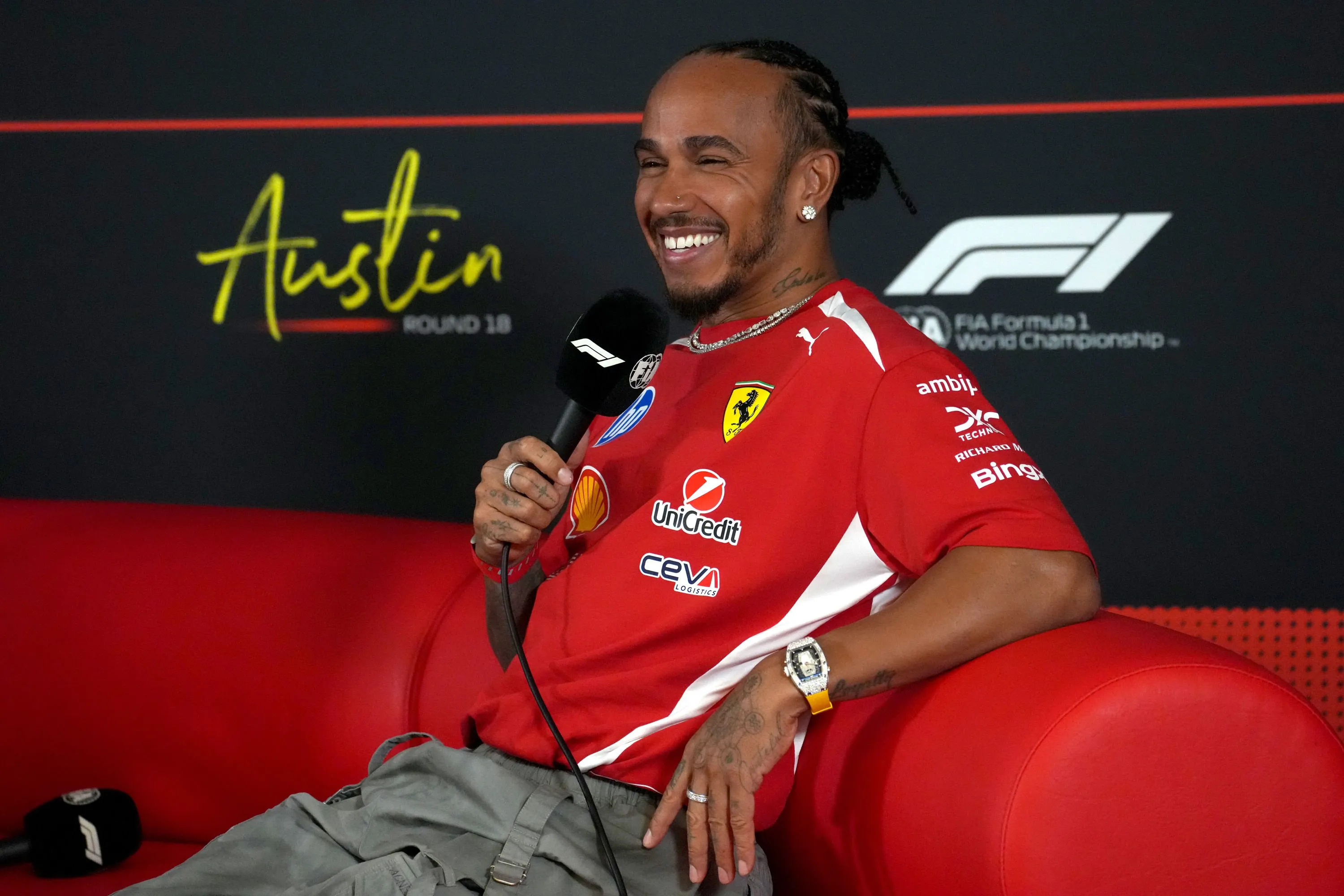 Lewis Hamilton smiling and holding a microphone while sitting on a red couch.