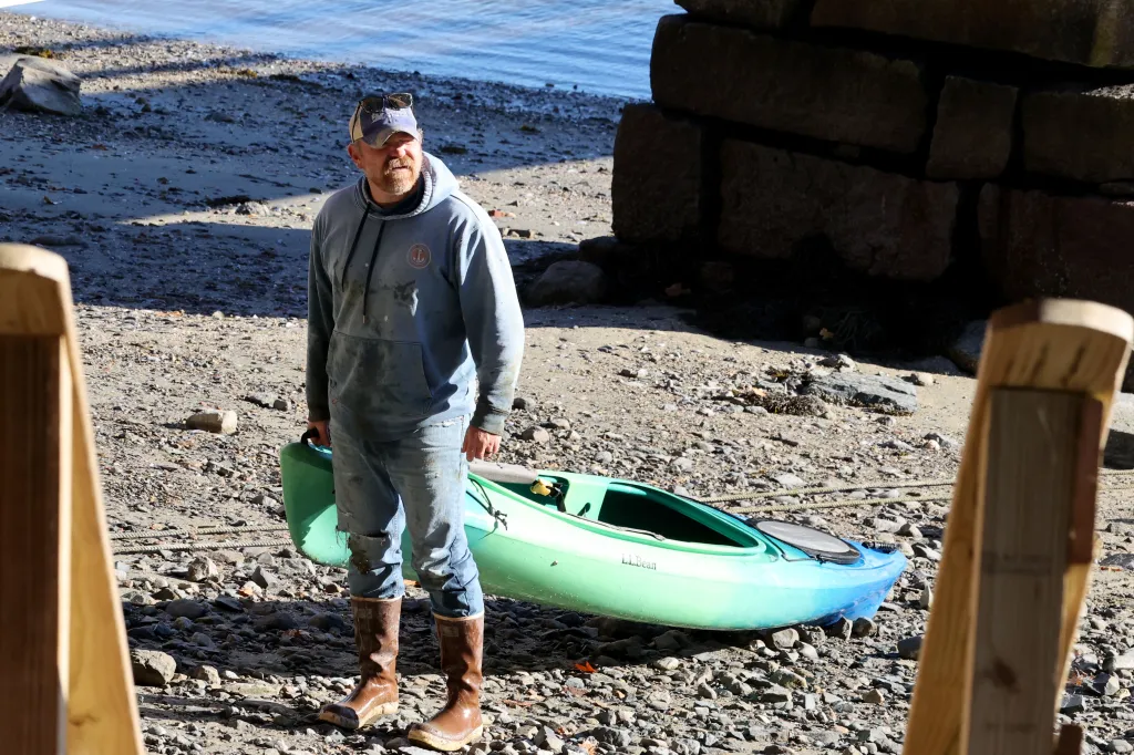 Graham Platner stands on a rocky shore next to a canoe.
