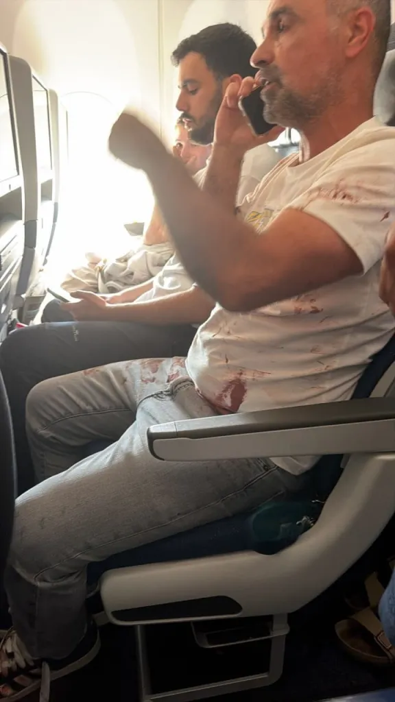 FlyDubai passenger Yaniv Hayun talking on the phone in an airplane seat, with blood stains visible on his white t-shirt and jeans after a cockpit attack.