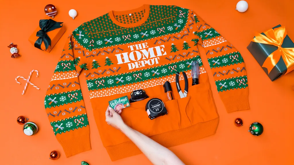 An orange and green holiday sweater featuring the The Home Depot logo, surrounded by wrapped gifts, ornaments, and tools tucked into its pockets.