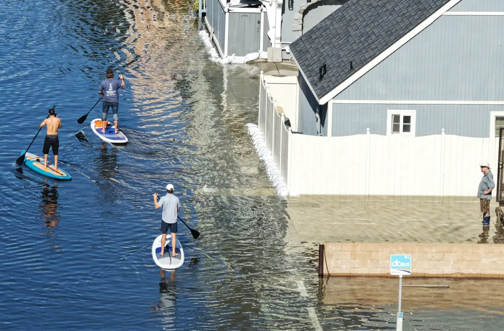 People paddle board in coastal floodwaters along a closed highway next to homes.
