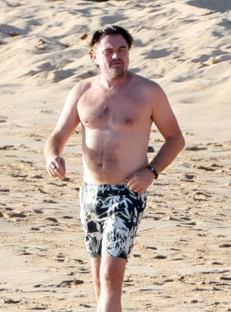 Leonardo DiCaprio walks shirtless along a sandy beach in swim trunks.