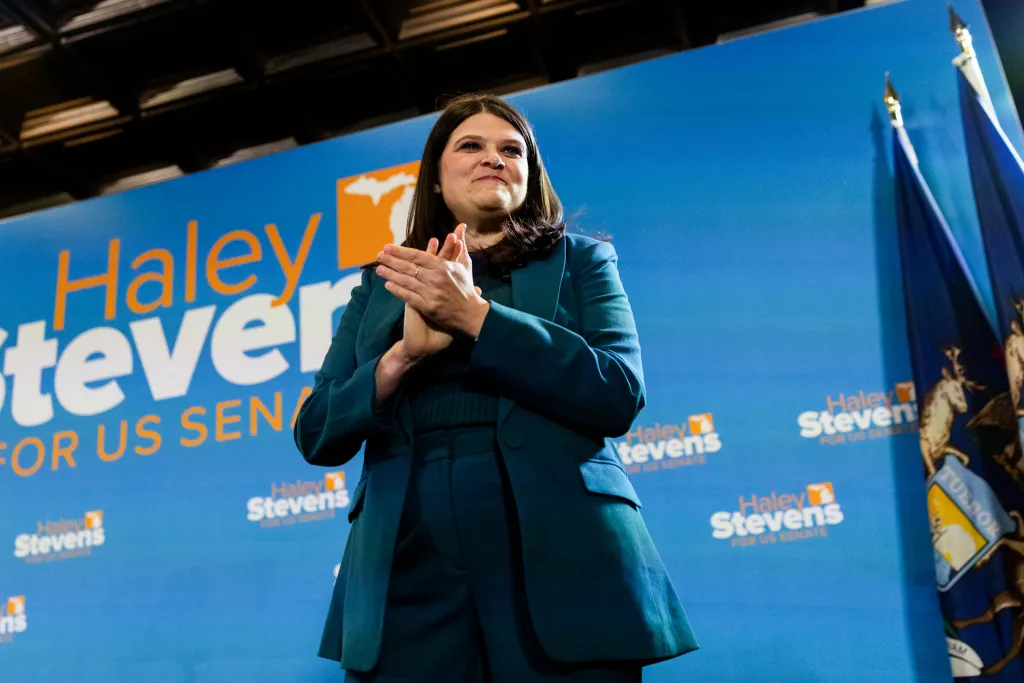 Rep. Haley Stevens applauds at an election night watch party in front of a blue backdrop bearing her campaign logo.