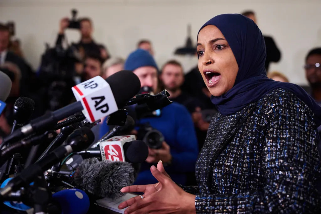 Rep. Ilhan Omar speaks at a news conference, surrounded by microphones and reporters.