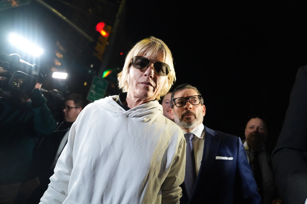 Selma Blair wearing sunglasses and a white hoodie, flanked by people at night.