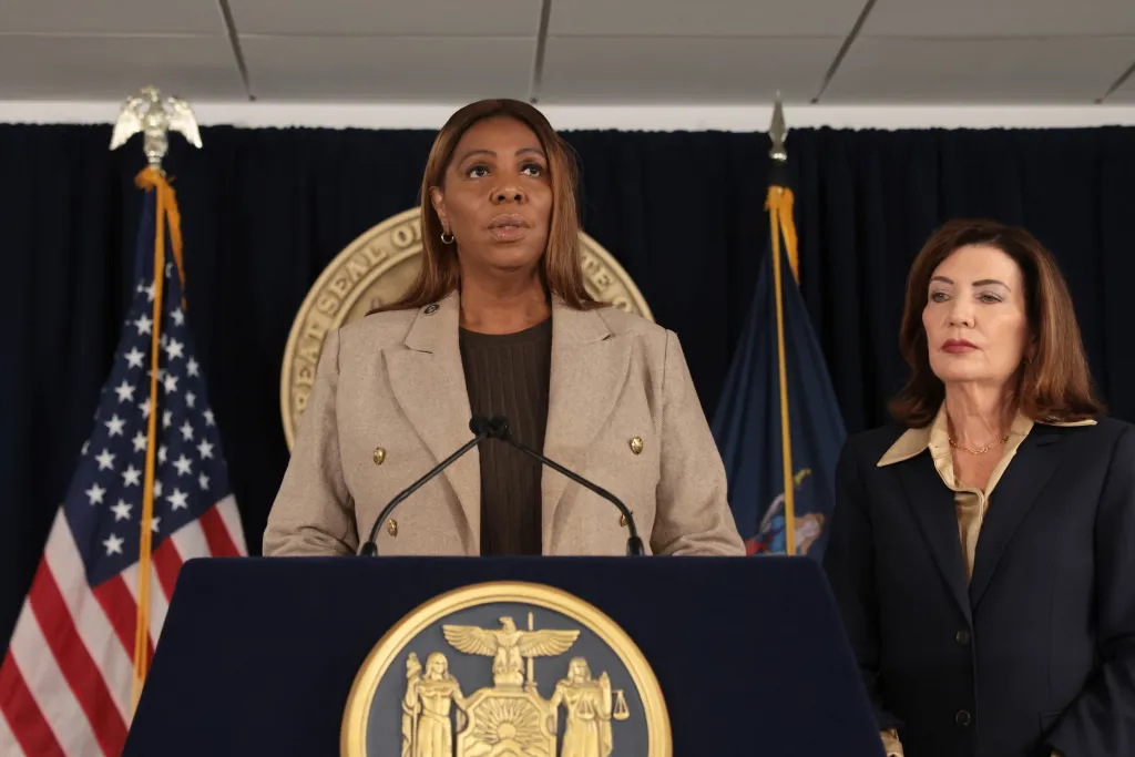 New York Attorney General Letitia James stands at a podium speaking, with New York Governor Kathy Hochul standing to her right.