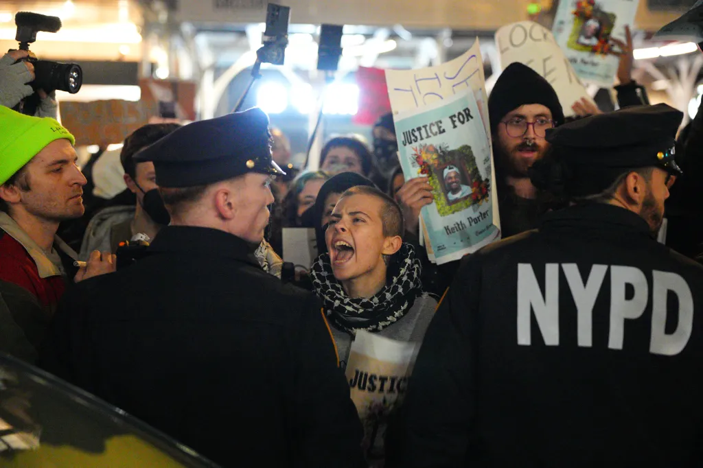 A protester with a shaved head and scarf shouts face-to-face with NYPD officers at Foley Square.