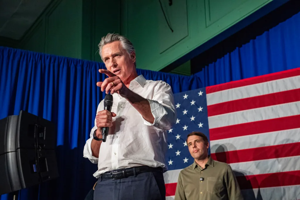 California Governor Gavin Newsom speaks while holding a microphone, with Democratic U.S. Senate candidate Chris Pappas standing in the background in front of an American flag.