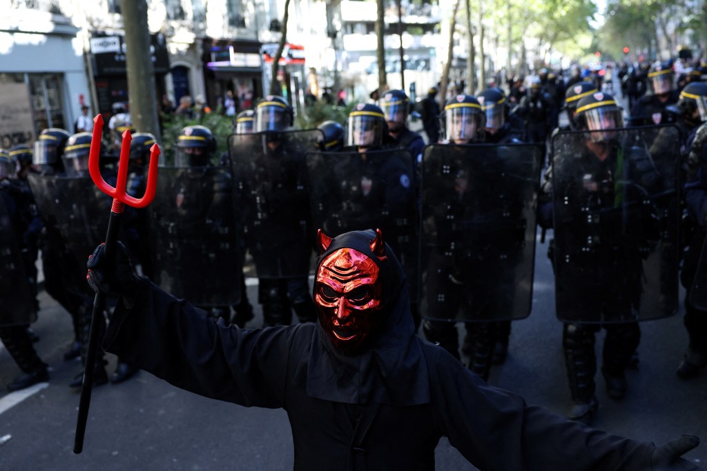 A protester in a black robe and red devil mask holds a red pitchfork in front of riot police during a student demonstration.