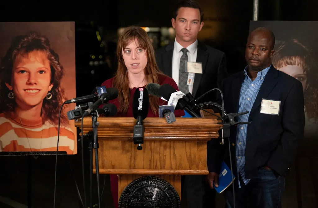 Witness Catherine Sweeney speaks at a podium with two men standing behind her and a poster of Colleen Slemmer beside her.