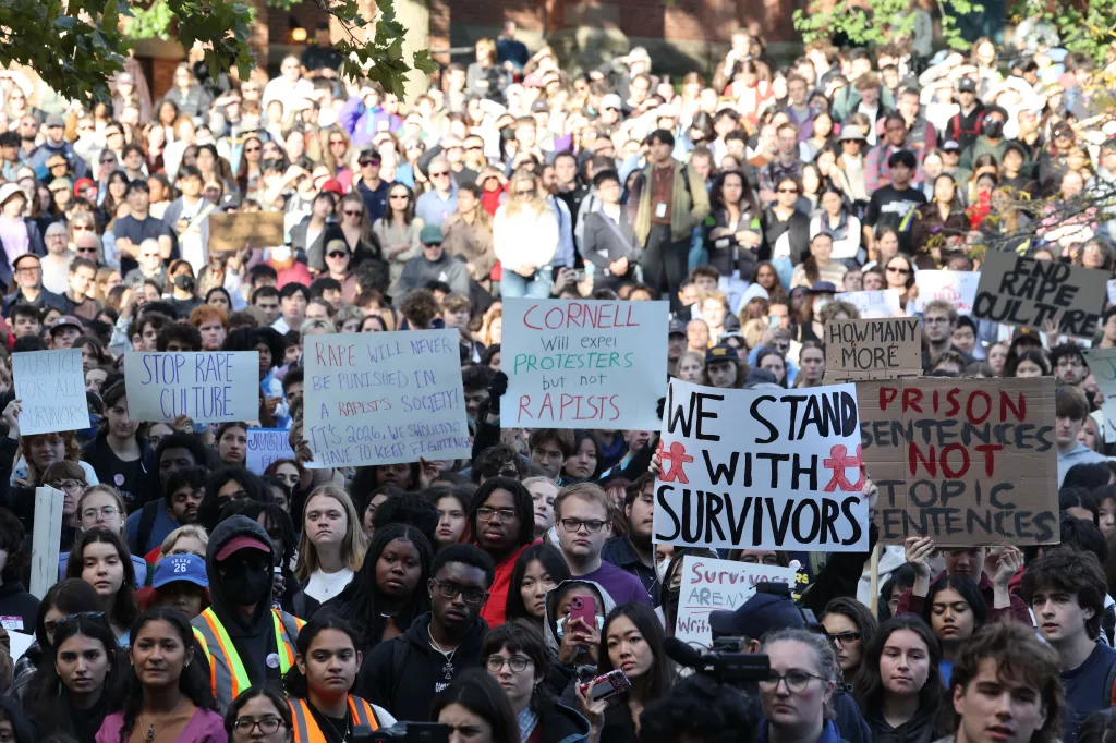 A large crowd of protesters gathers on a campus, holding signs with messages like 
