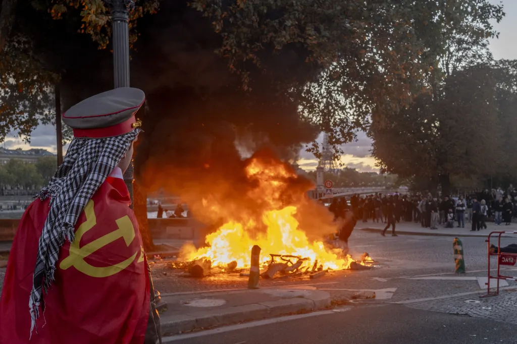 A protester draped in a communist flag watches a street fire during a cost of living demonstration in Paris.