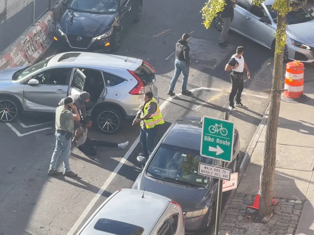 A man is detained on the ground by law enforcement officers next to a silver SUV on a New York City street.