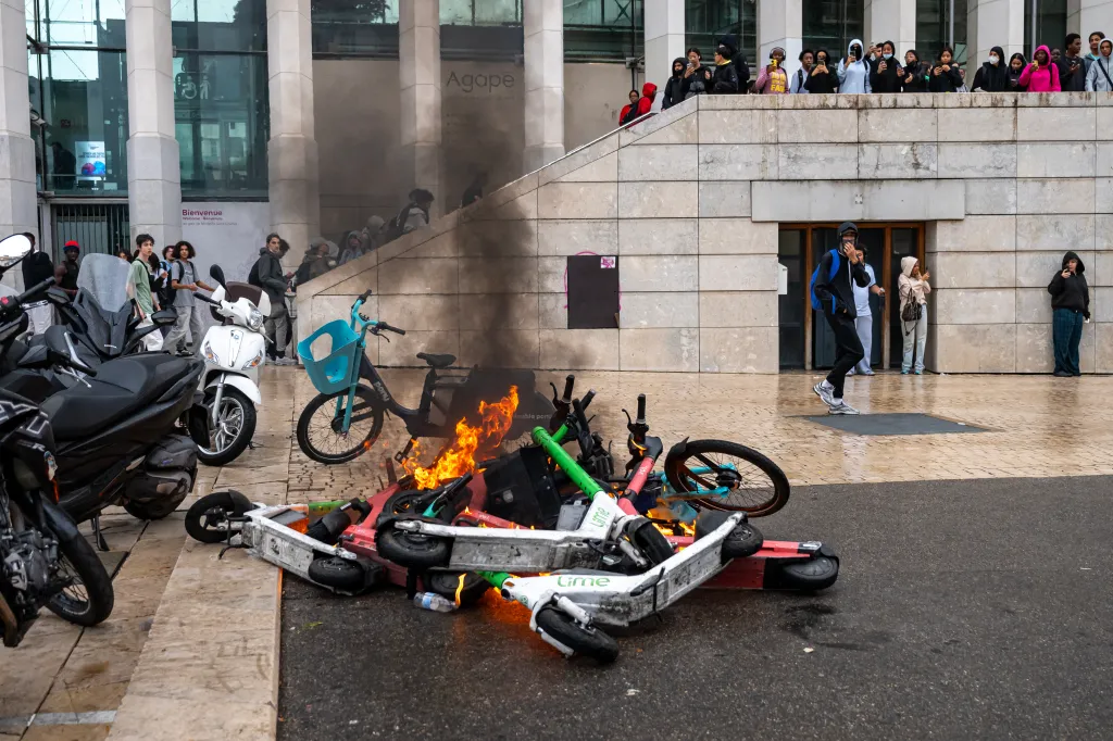 A pile of burning e-scooters smokes on the ground outside a building, with a crowd of students watching from behind.