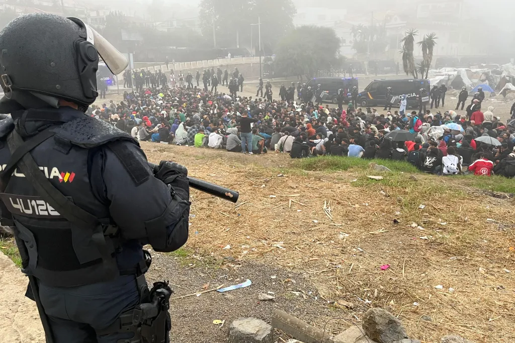 National Police officer in riot gear looks over a large group of migrants gathered near a beach camp.