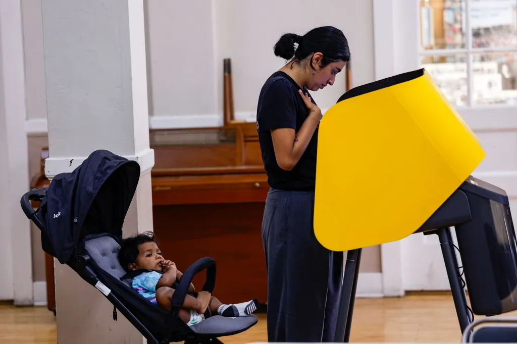 A woman casts her ballot at a voting machine while a young child sits in a stroller next to her.
