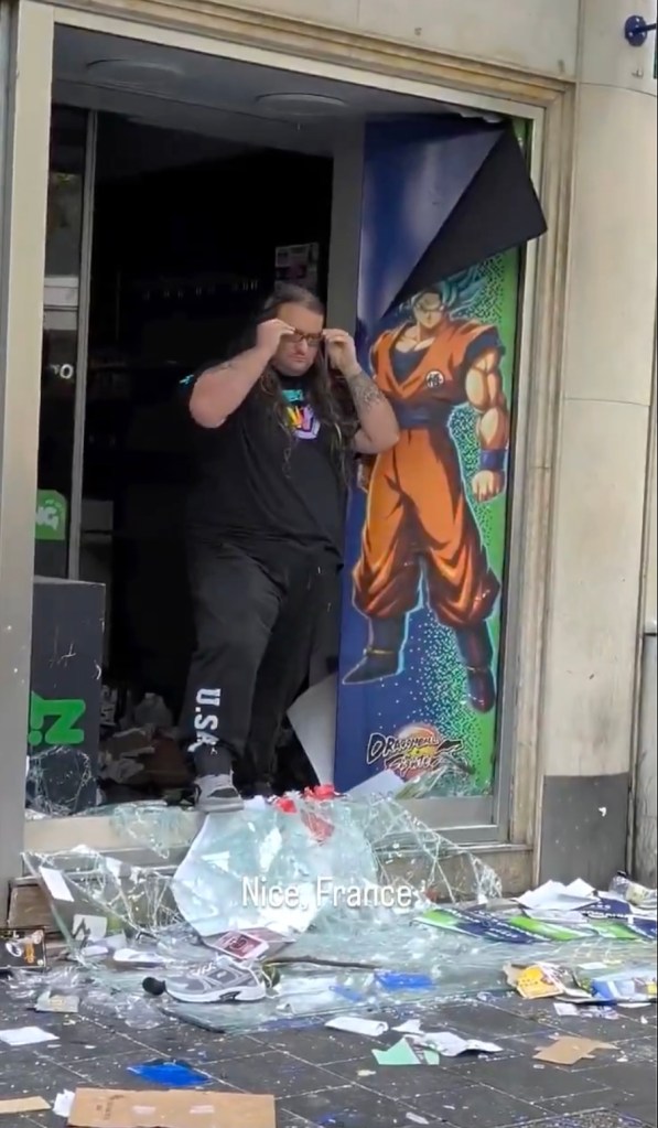 Pierre, a sales assistant, stands in the shattered doorway of his video game store in Nice, France, next to a peeled poster of Goku.