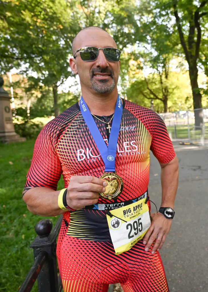 Lee Anton proudly displays his finisher's medal after completing the Big Apple Triathlon, wearing a red and black race suit and sunglasses.