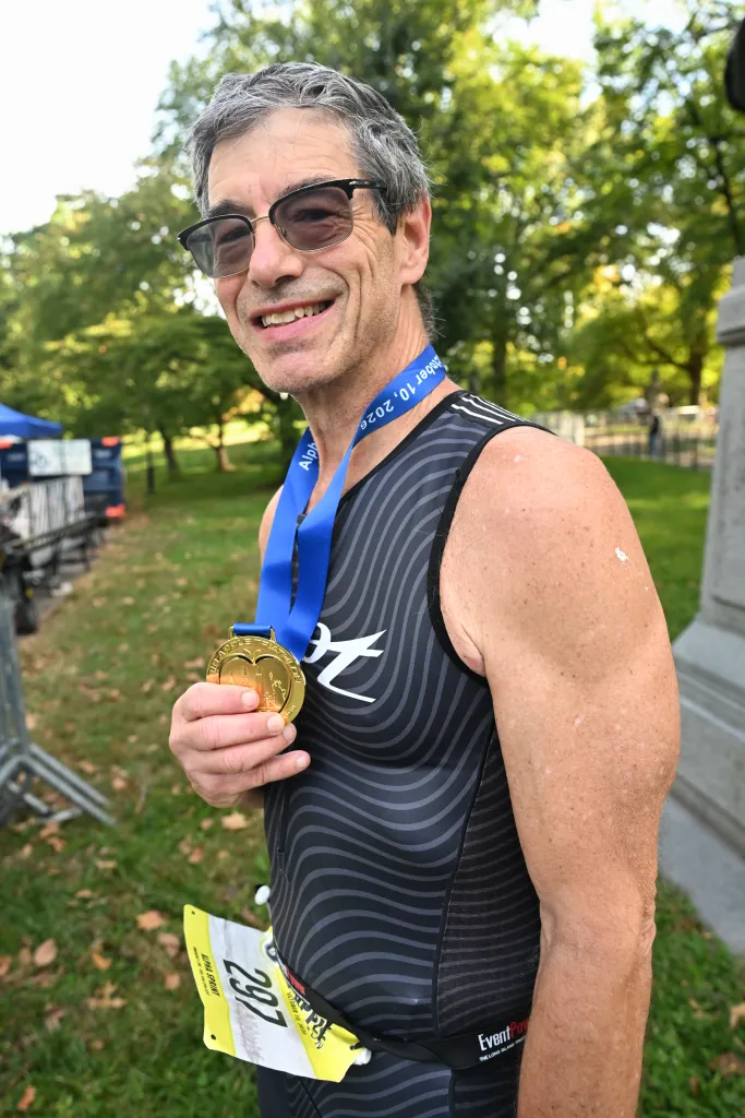 Jack Hershman, 71, wearing a triathlon race suit and sunglasses, proudly holds a gold medal after finishing first in his age group at the Big Apple Triathlon.