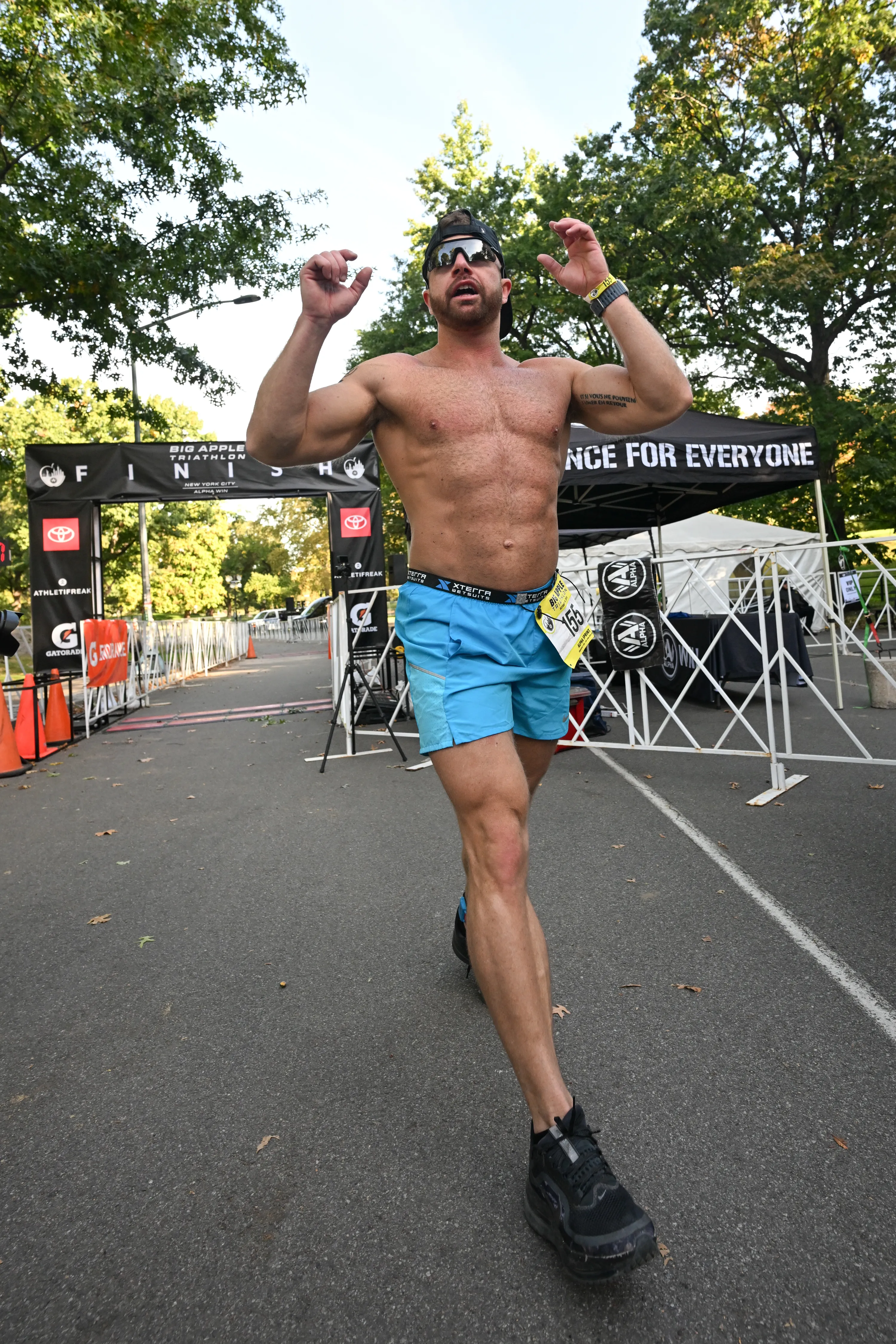 A shirtless male athlete wearing sunglasses and blue shorts celebrates as he crosses the finish line at the Big Apple Triathlon.