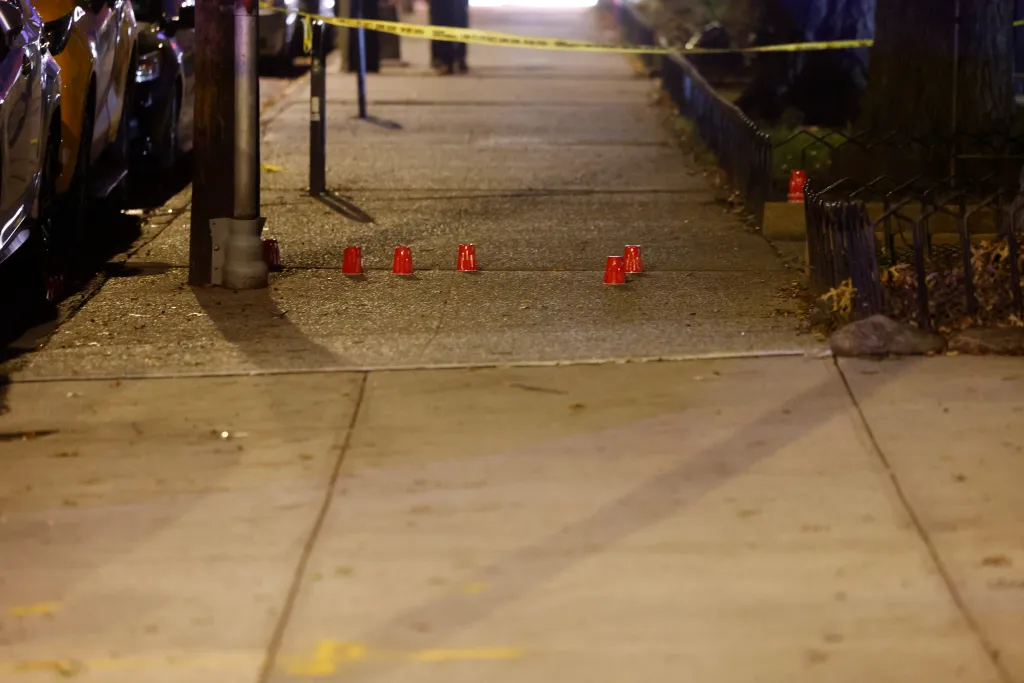 Police investigate a shooting scene on a sidewalk in the Bronx, marked by red cups and yellow tape.
