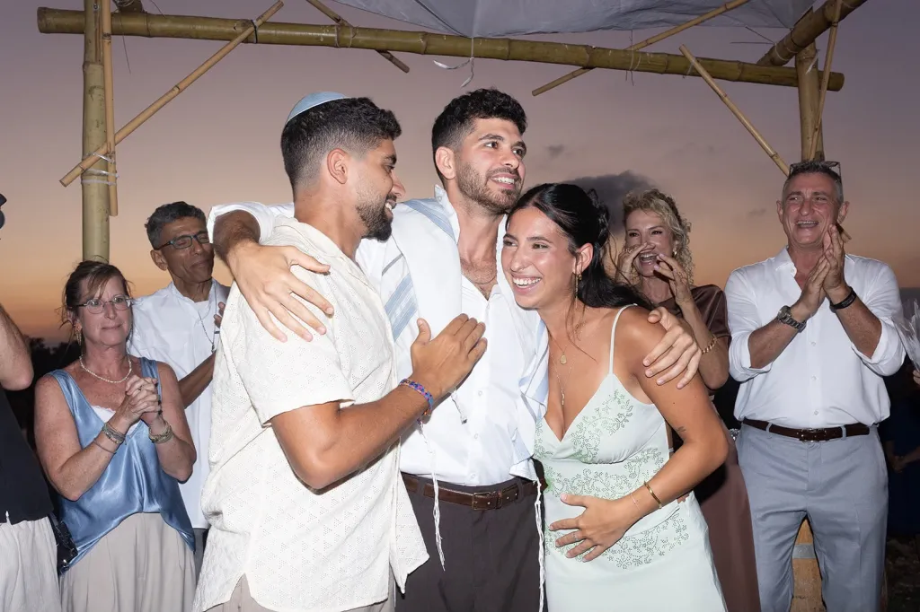 The David siblings, Evyatar, Ilay, and Yeela, embrace and smile as onlookers applaud in the background.