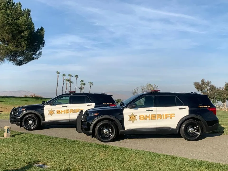 Two black and white Riverside County Sheriff's patrol cars parked on a grassy field.