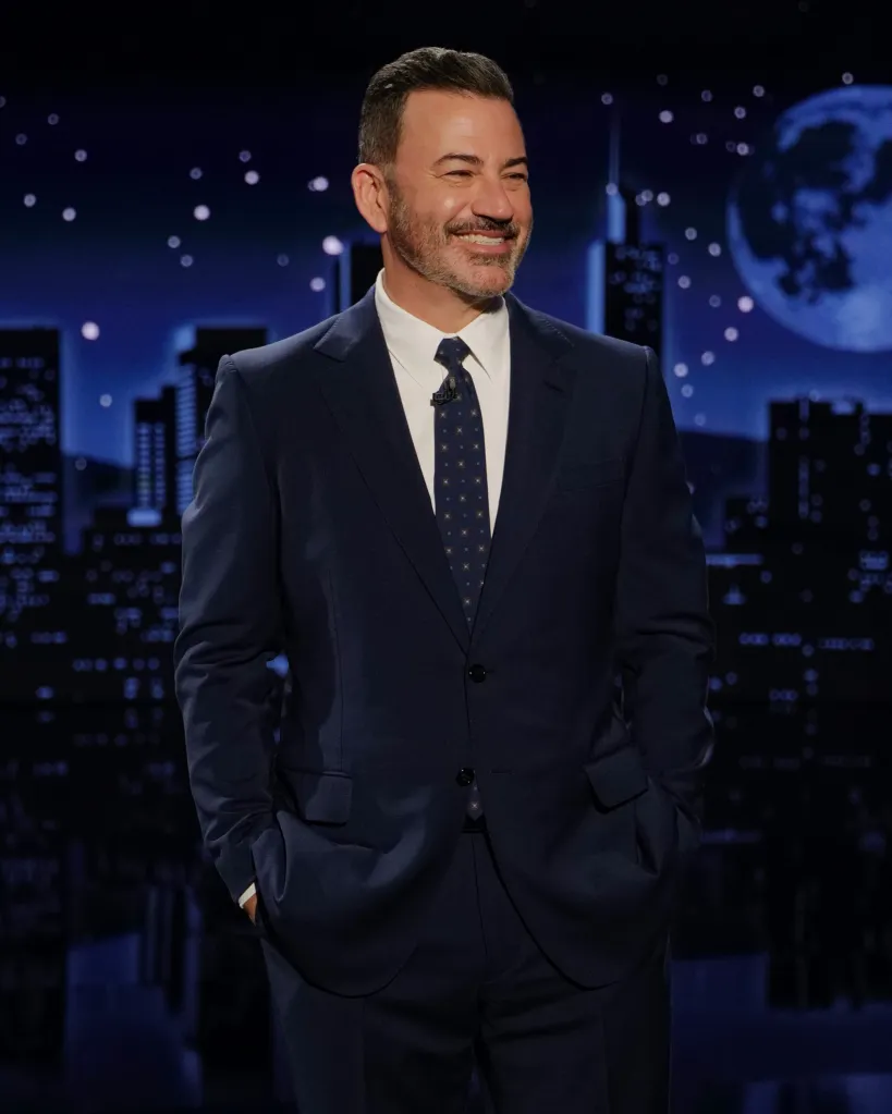 Jimmy Kimmel smiling in a suit on stage with a city skyline background.