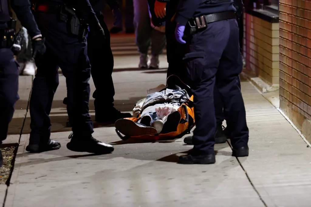 Police officers and emergency personnel respond to a shooting and barricaded suspect situation, standing over a victim on a stretcher on a sidewalk.