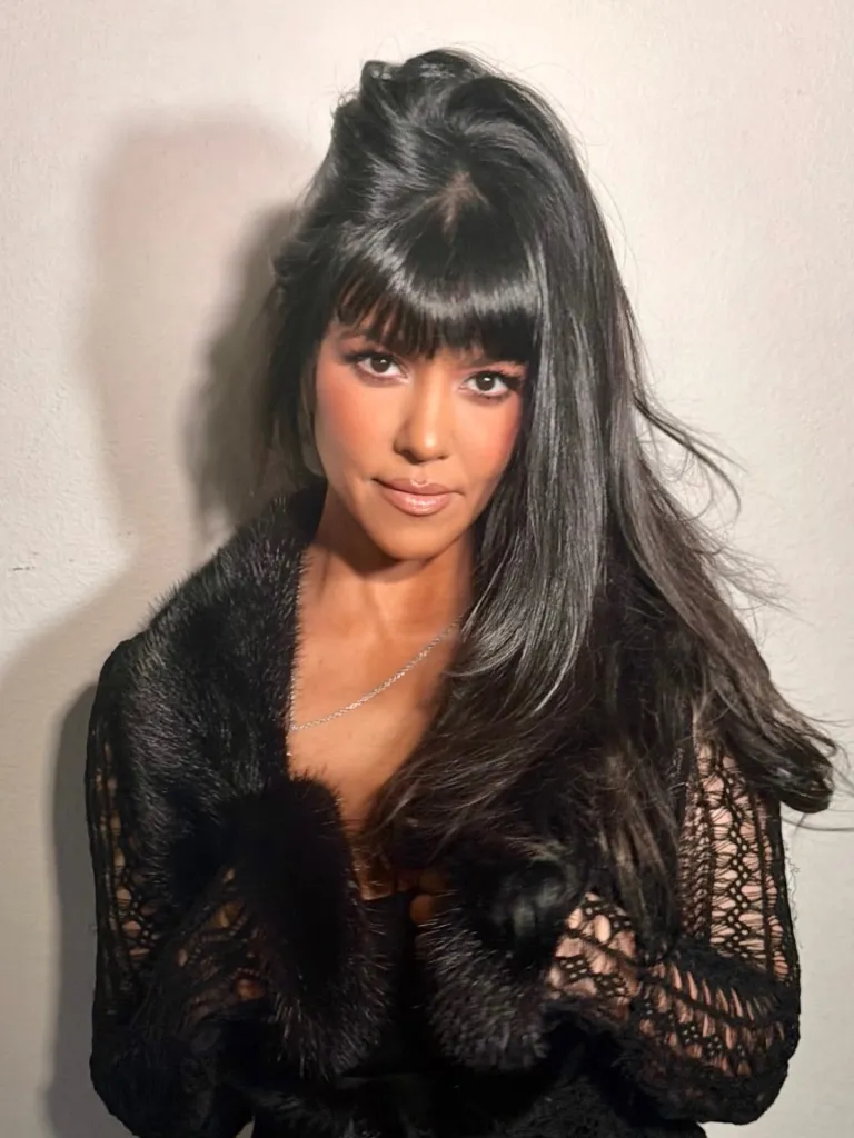 Kourtney Kardashian with long dark hair, bangs, and a black lace and fur garment.