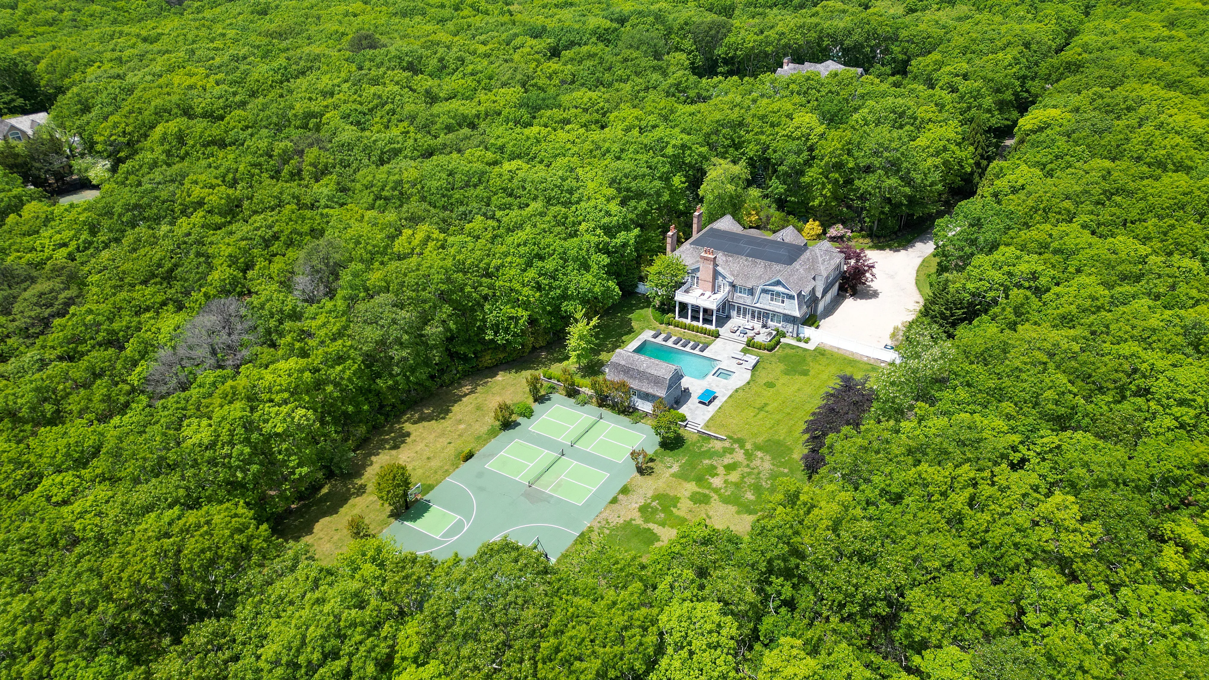 Aerial view of Libbie Mugrabi's home in Bridgehampton surrounded by dense trees, featuring a tennis court, swimming pool, and a large house.