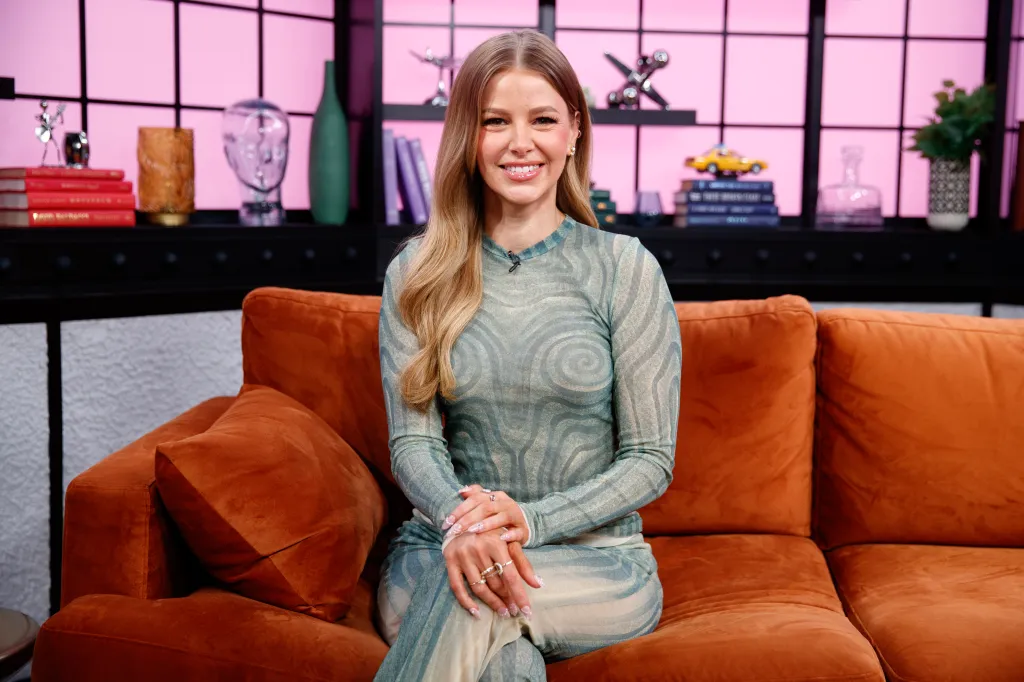 Ariana Madix sits on a rust-colored couch during an interview in a pink-walled studio with shelves holding books, a green vase, and other decorative items.