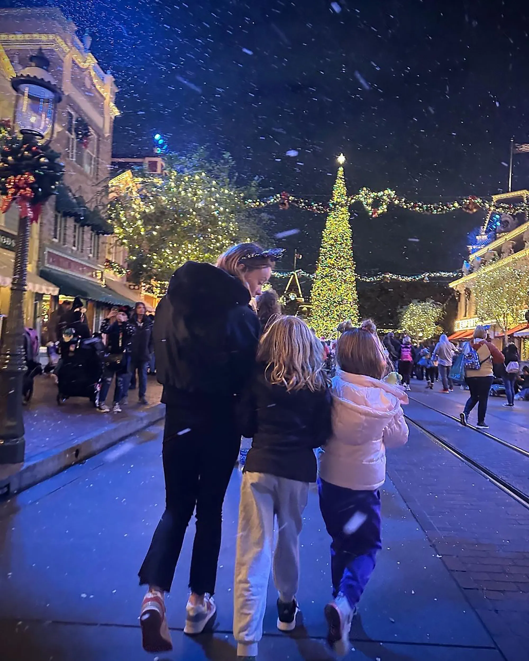 A woman and two young girls walk together down a crowded street lit by festive decorations and a large Christmas tree, as snow falls.