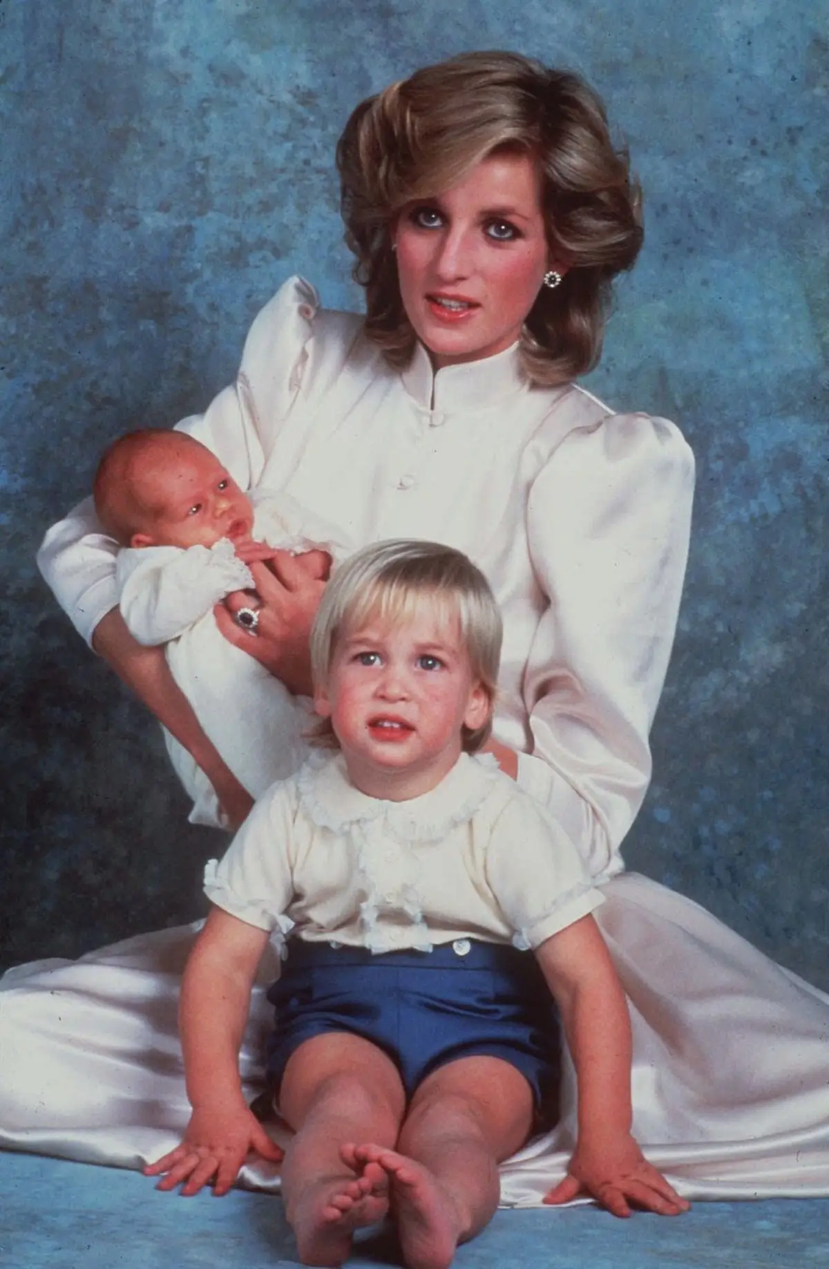Princess Diana holds a newborn Prince Harry in her arms while Prince William looks on.