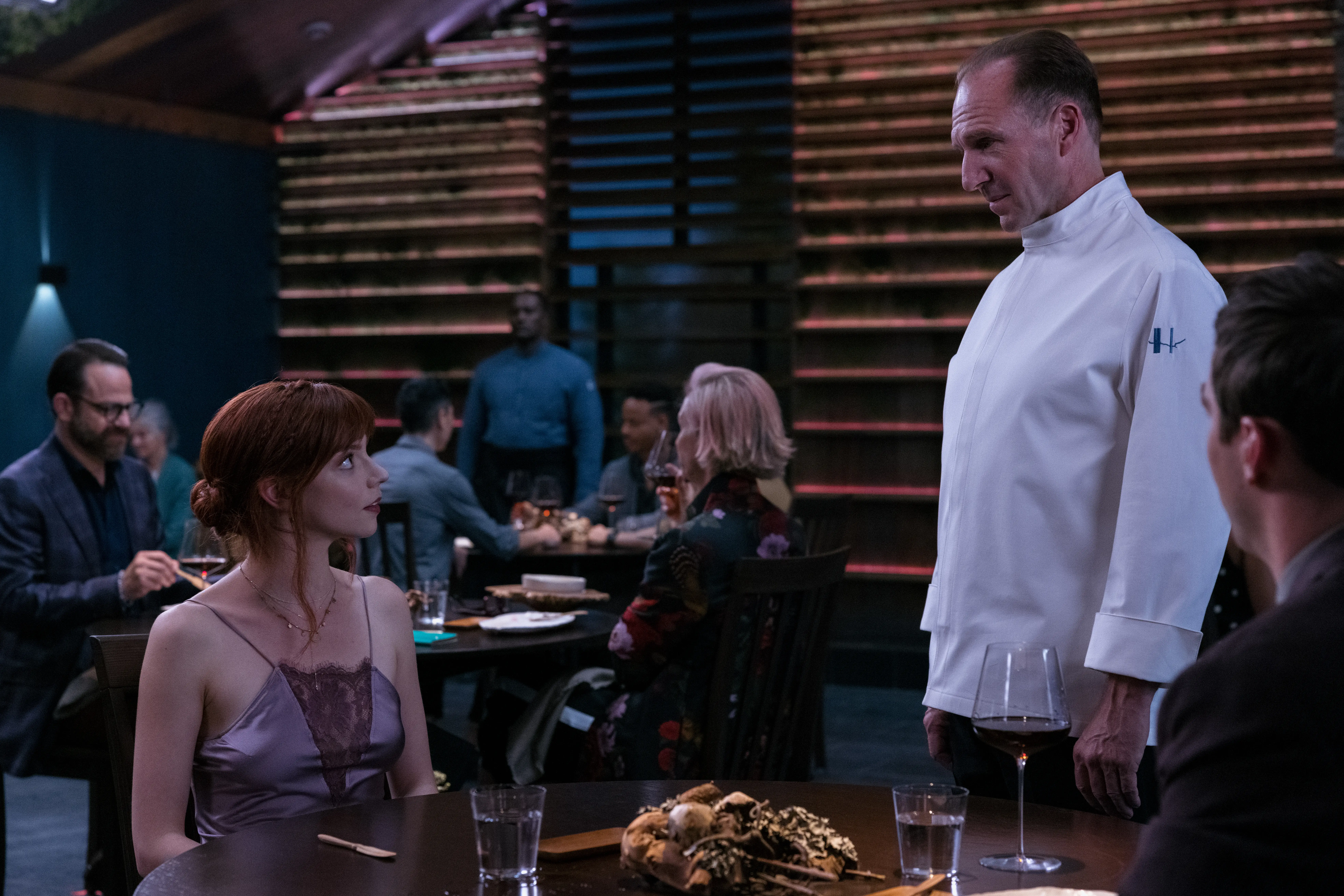 Anya Taylor-Joy and Ralph Fiennes in The Menu with one seated and the other standingin a restaurant