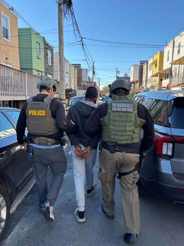 Destino Jean Louis in handcuffs being escorted down a city street by a police officer and a U.S. Marshal.