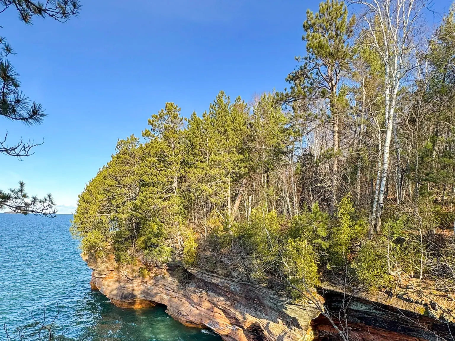 Emily sits on the ground in front of a blue-green body of water and trees.