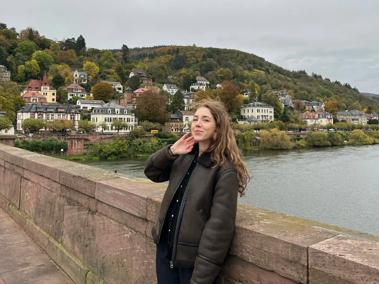 Dasha leans against a bridge over a river. Behind her are hills with houses on them.