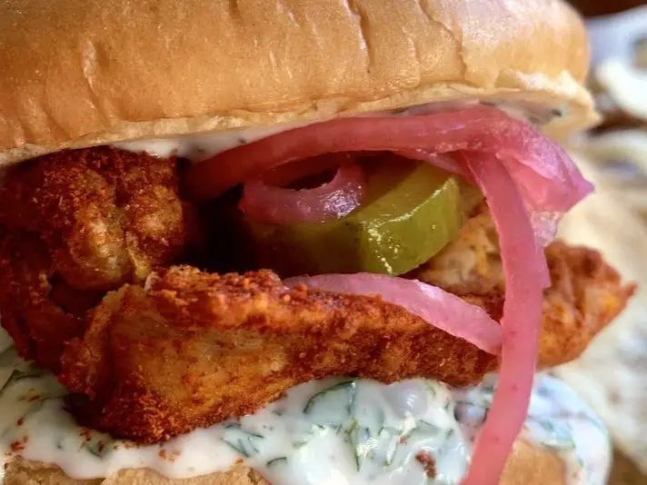 A close-up of a chicken sandwich with pink pickled onions, pickles, and sauce.