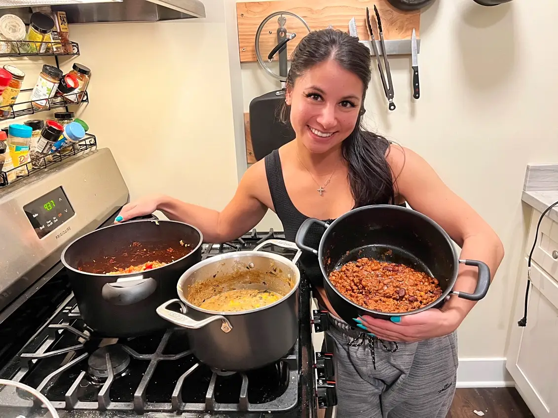 The writer Chelsea Davis stands next to a stove and holds a black pot of a chili and puts her arm around two other pots of chili