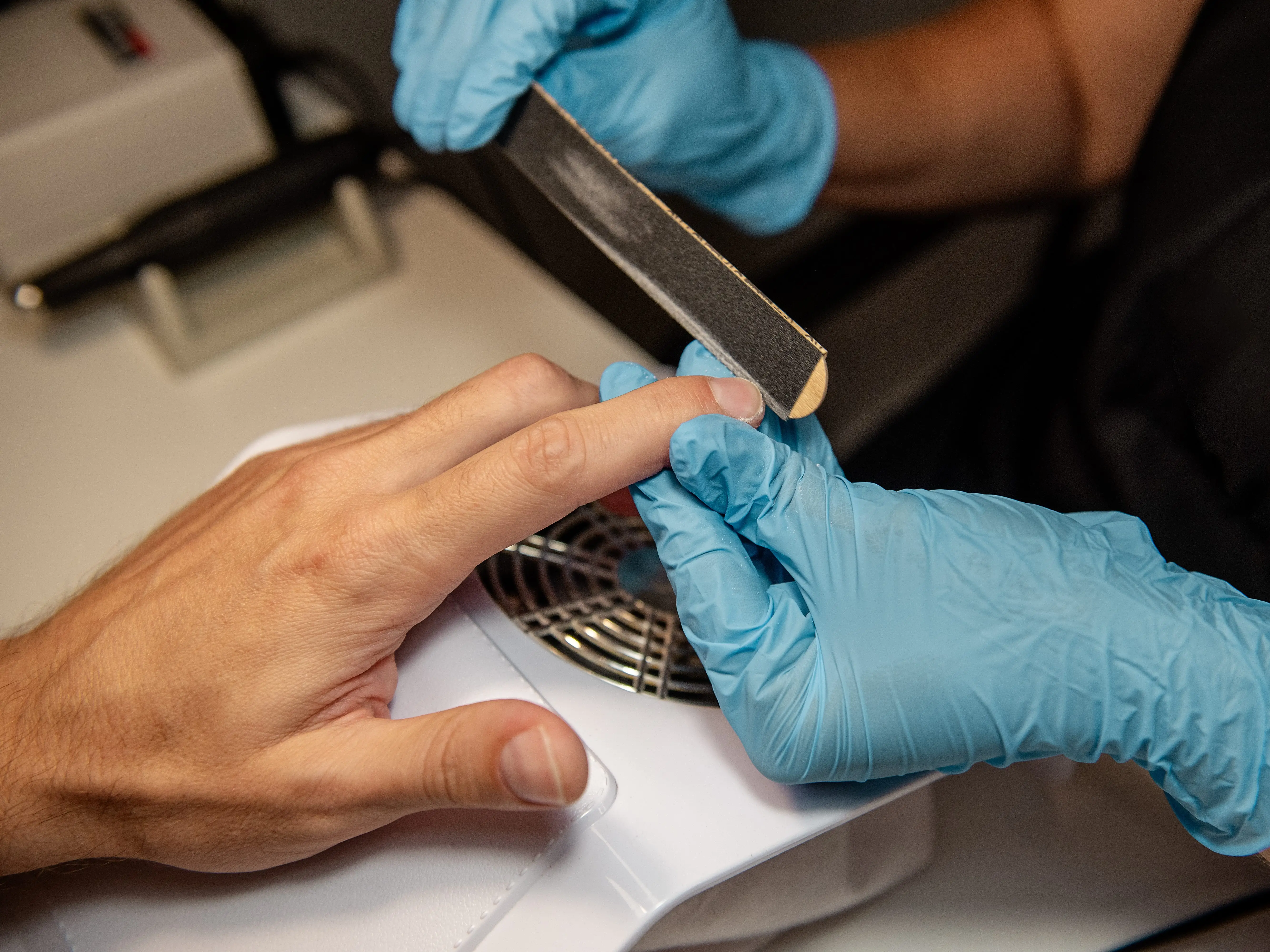A close-up of a nail technician with blue gloves filing a man's hand at a salon.